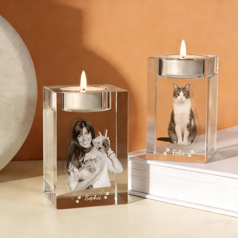 May include: Two clear glass candle holders with lit candles. One features a woman with a dog, the other a cat. The woman's holder has the name "Sophie" and paw prints. The cat's holder has the name "Felix" and paw prints.