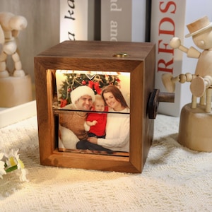 Könnte beinhalten: Ein Holz-Fotowürfel mit einem beleuchteten Familienfoto. Der Würfel hat einen Drehmechanismus an der Seite und einen kleinen Messingknopf oben. Das Familienfoto zeigt einen Mann, eine Frau und ein Kind vor einem Weihnachtsbaum.