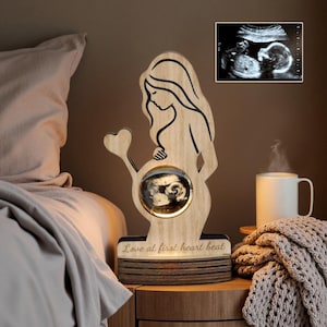 May include: Wooden pregnancy keepsake with a pregnant person silhouette holding a heart, and a sonogram image in the center. The base reads "Love at first heart beat". A sonogram image is in the upper right corner.
