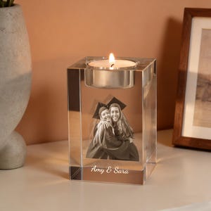 May include: A clear, square glass candle holder with a lit tea light. The holder features a black and white photo of two people in graduation attire, with the names "Amy & Sara" below the photo. Home decor.