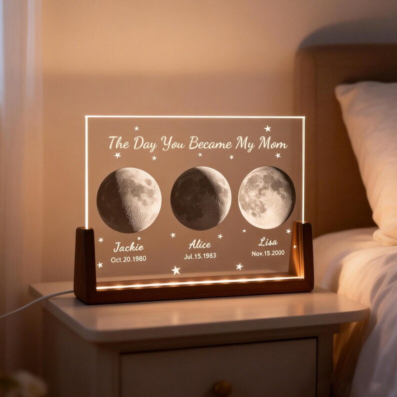 May include: A personalized night light with a wooden base and a clear acrylic panel. The panel features three moon phases, names, and dates, with the text "The Day You Became My Mom". The light is on a white nightstand.