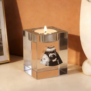 May include: A clear, square crystal candle holder with a lit candle. An ultrasound image is displayed on the front of the holder. The candle holder is on a light-colored surface.