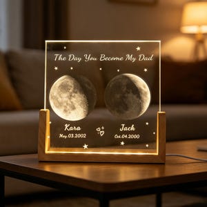 Puede incluir: Una luz nocturna rectangular de acrílico con base de madera clara, que muestra dos fases lunares iluminadas, el texto "The Day You Become My Dad", los nombres "Kara" y "Jack", y las fechas "May.03.2002" y "Oct.04.2000" con estrellas y corazones.