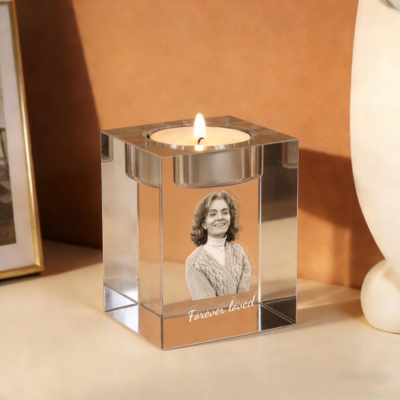 May include: A clear, square crystal candle holder with a lit candle. The holder features a photo of a woman and the text "Forever loved". The candle holder is on a wooden surface.