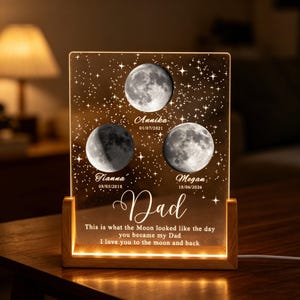 Pode incluir: Uma luz noturna retangular de acrílico com base de madeira exibindo três fases da lua, os nomes Annika, Tianna e Megan com datas, e o texto: Dad, This is what the Moon looked like the day you became my Dad, I love you to the moon and back.
