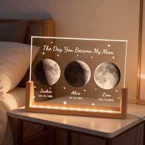 May include: A personalized night light with a wooden base and a clear acrylic panel. The panel features three moon phase images, names, and dates, with the text "The Day You Became My Mom" and star accents. The light is on a white table.