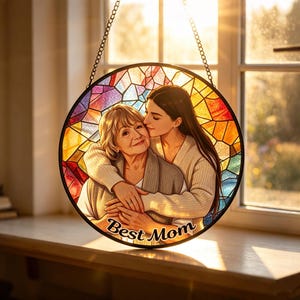 Pode incluir: Arte de vitral redondo com uma mãe e filha se abraçando, com o texto "Best Mom" na parte inferior. O fundo é um padrão de mosaico colorido. A janela é suspensa por uma corrente.