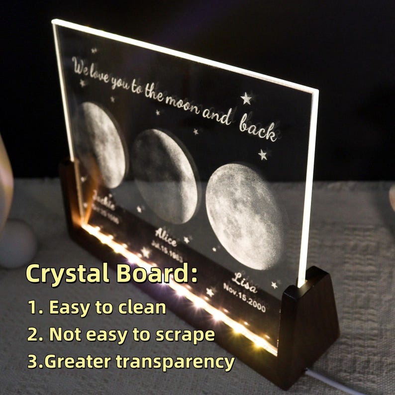 May include: A personalized crystal board with a dark background, featuring moon phases and the text "We love you to the moon and back." Names and dates are inscribed below the moon phases. The board is illuminated by warm white LED lights.