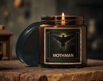 May include: A lit soy wax candle in a brown glass jar with a black label featuring a Mothman graphic. The jar has a black lid and sits on a wooden surface. The text "Mothman" is visible on the label.