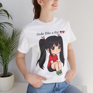 May include: White t-shirt featuring a cartoon girl in a red top and green shoes. The text "Code Like a Girl" is printed on the shirt with a red heart and a laptop icon.