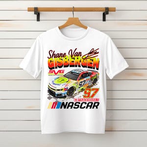 Pode incluir: Camiseta branca com um gráfico colorido de um carro de corrida com o número 97 e o texto "Shane Van Gisbergen" e "NASCAR". O design inclui o logotipo da Trackhouse.