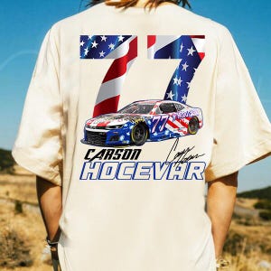 Op de afbeelding: Cr&egrave;mekleurig T-shirt met een afbeelding van een raceauto, het nummer 77 en de naam "Carson Hocevar". Het ontwerp bevat de sterren en strepen van de Amerikaanse vlag. De auto is blauw, rood en wit.