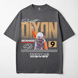 May include: Dark gray t-shirt featuring a graphic of IndyCar driver Scott Dixon. The design includes the name "Scott Dixon" in a stylized font, the number "9", and the words "INDYCAR DRIVER". The shirt has a crew neck and short sleeves.
