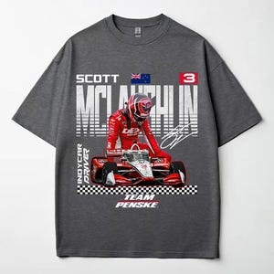 May include: Dark gray t-shirt featuring a graphic of a race car and driver in a red racing suit. The shirt includes the text "SCOTT McLAUGHLIN" and "INDYCAR DRIVER" with the Team Penske logo.