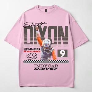 May include: A light pink t-shirt featuring a graphic of IndyCar driver Scott Dixon. The design includes the driver's name, the number 9, and the words "INDYCAR DRIVER". The shirt also features a checkered flag design.
