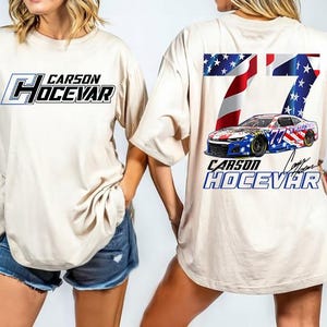 Op de afbeelding: Beige T-shirt met de naam "CARSON HOCEVAR" in zwarte en blauwe letters op de voorkant. De achterkant toont het nummer "77" met een Amerikaans vlagontwerp en een raceauto-afbeelding. Het shirt heeft een casual, oversized pasvorm.