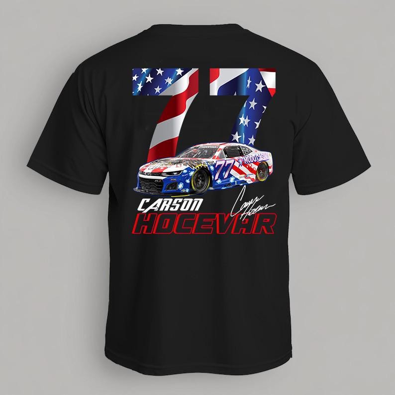 Op de afbeelding: Zwart T-shirt met een afbeelding van een raceauto, nummer 77 en een ontwerp van de Amerikaanse vlag. Het shirt toont ook de naam "Carson Hocevar" in rode en witte tekst.