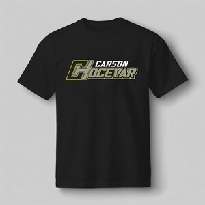 Op de afbeelding: Zwart T-shirt met de naam "CARSON HOCEVAR" in een gestileerd lettertype. De tekst is voornamelijk wit met gele en grijze accenten. Het T-shirt heeft een ronde hals en korte mouwen.