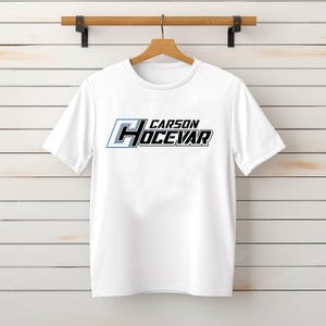 Op de afbeelding: Wit T-shirt met de tekst "CARSON HOCEVAR" in zwarte en blauwe letters. Het T-shirt hangt aan een houten hanger tegen een wit geschilderde houten plankmuur.