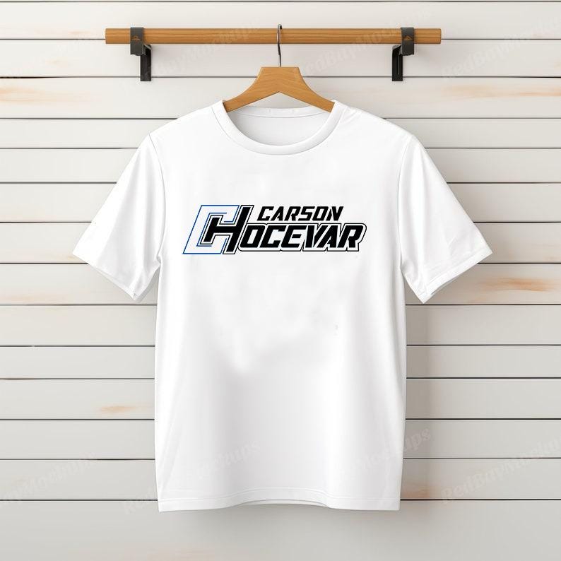 Op de afbeelding: Wit T-shirt met de tekst "CARSON HOCEVAR" in zwarte en blauwe letters. Het T-shirt hangt aan een houten hanger tegen een wit geschilderde houten plankmuur.
