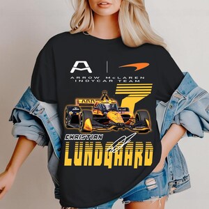 May include: Black t-shirt featuring a graphic of an IndyCar race car in yellow and black, with the text "ARROW MCLAREN INDYCAR TEAM" and the name "CHRISTIAN LUNDGAARD". The shirt also displays the number 7 and a signature.