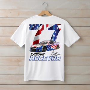 Op de afbeelding: Wit t-shirt met een raceauto-ontwerp, het nummer 77 en de naam "Carson Hocevar". Het ontwerp bevat de sterren en strepen van de Amerikaanse vlag. Het t-shirt hangt aan een houten hanger.