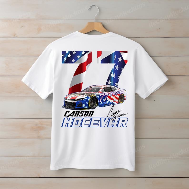 Op de afbeelding: Wit t-shirt met een raceauto-ontwerp, het nummer 77 en de naam "Carson Hocevar". Het ontwerp bevat de sterren en strepen van de Amerikaanse vlag. Het t-shirt hangt aan een houten hanger.