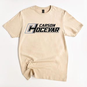 Op de afbeelding: Een beige t-shirt met de naam "CARSON HOCEVAR" in een opvallend, sportief lettertype over de borst. De tekst is voornamelijk zwart met blauwe en witte accenten. Het t-shirt is gemaakt van een zacht materiaal.