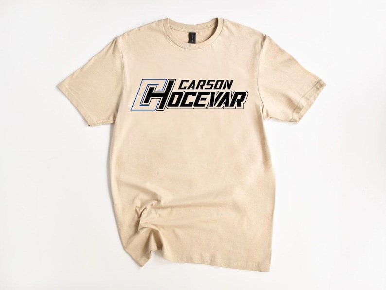 Op de afbeelding: Een beige t-shirt met de naam "CARSON HOCEVAR" in een opvallend, sportief lettertype over de borst. De tekst is voornamelijk zwart met blauwe en witte accenten. Het t-shirt is gemaakt van een zacht materiaal.