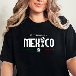 May include: Black t-shirt featuring the text "MEXICO" in bold white letters, with a soccer trophy graphic. The shirt also includes the text "SELECCIÓN NACIONAL DE" above the word "MEXICO" and a green, white, and red stripe design.