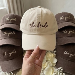 May include: A collection of baseball caps, with one cream-colored cap embroidered with "the bride" in brown script, surrounded by several brown caps embroidered with "the girls" in white script. The caps are arranged on a surface with floral accents.