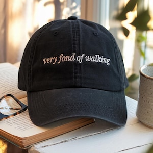 May include: Black baseball cap with the embroidered text "very fond of walking" in white. The cap is sitting on a white surface next to an open book and a mug.