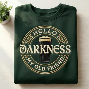May include: Dark green sweatshirt with a circular embroidered design. The design features a pint of dark beer with the words "HELLO DARKNESS MY OLD FRIEND" in a gold color. The sweatshirt is folded.
