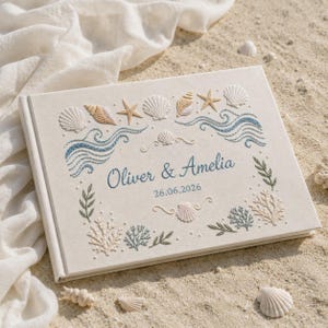 May include: A rectangular off-white guest book featuring embroidered coastal motifs including seashells, starfish, and blue wave patterns. The center displays the text Oliver & Amelia and the date 26.06.2026 in blue embroidery, surrounded by coral and seaweed designs.