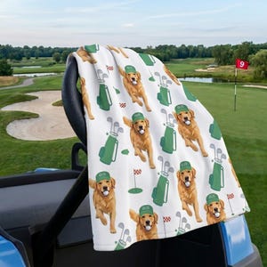 May include: White golf towel featuring a repeating pattern of a golden retriever wearing a green golf hat and holding a golf ball. The towel also includes green golf bags, golf clubs, and golf flags. The word "GUS" is on the hat.