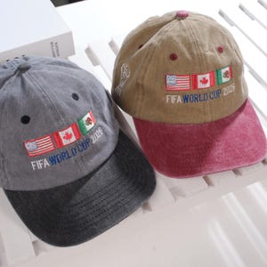 May include: Two baseball caps. One is gray with a black brim, the other is tan with a maroon brim. Both caps have the text "FIFA WORLD CUP 2026" embroidered below the flags of the United States, Canada, and Mexico.