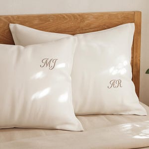 May include: Two white square pillows with embroidered initials, "MJ" and "AR", rest on a bed. The pillows are made of a smooth, light-colored fabric and are placed against a wooden headboard. The overall aesthetic is simple and elegant.
