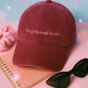 May include: A maroon baseball cap with the embroidered text "hot girls read books." The cap is displayed with a pink scrunchie, a spiral-bound notebook, and black cat-eye sunglasses. The items are arranged on a pink surface.