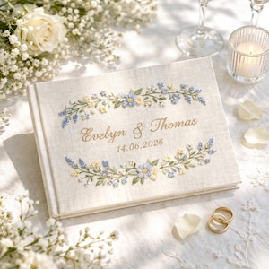 May include: A rectangular guest book with a beige linen cover featuring embroidered blue and yellow floral garlands. The center displays the gold embroidered text Evelyn & Thomas and the date 14.06.2026 in a script and serif font.