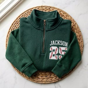 May include: A dark green pullover sweatshirt with a quarter-zip neckline. The sweatshirt features the name "JACKSON" and the number "25" in a baseball-themed design on the front. The sweatshirt is displayed on a woven, circular mat.