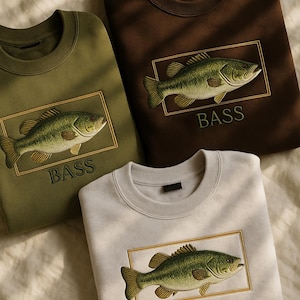 May include: Three crewneck sweatshirts in olive green, brown, and light gray. Each features an embroidered bass fish design within a gold rectangular frame, with the word "BASS" below. The sweatshirts are displayed on a textured, off-white surface.