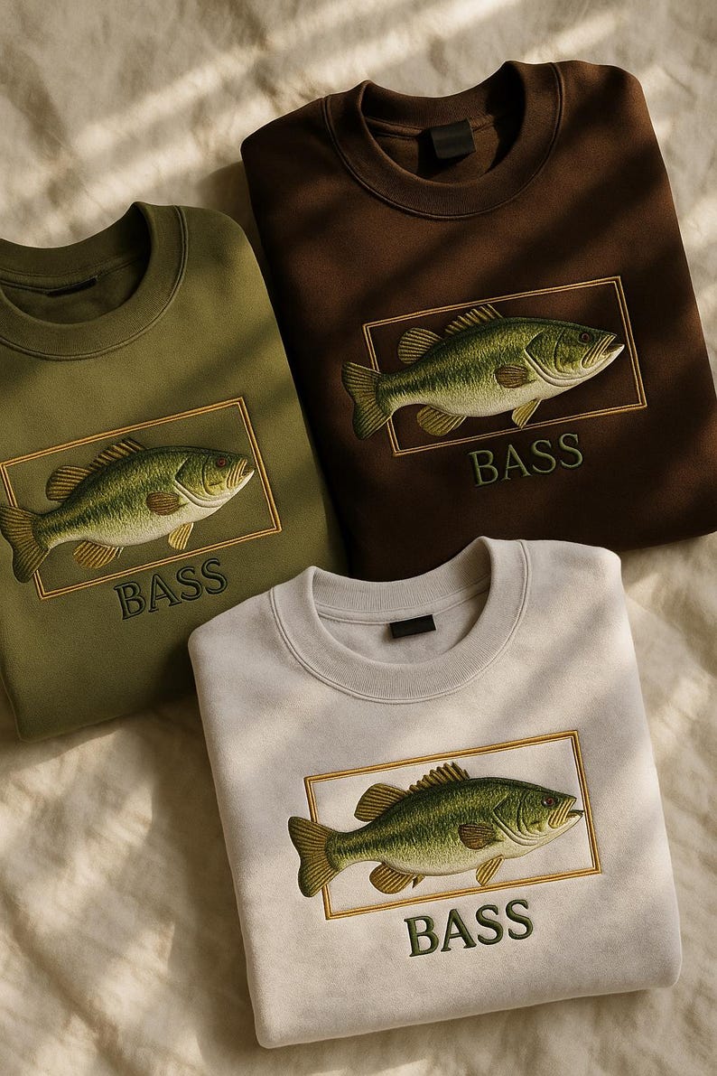 May include: Three crewneck sweatshirts in olive green, brown, and light gray. Each features an embroidered bass fish design within a gold rectangular frame, with the word "BASS" below. The sweatshirts are displayed on a textured, off-white surface.