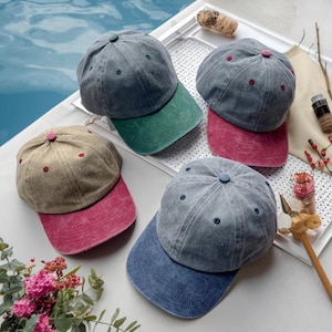 May include: A collection of baseball caps in various colors, including gray, green, brown, and blue. Each cap features a contrasting color on the brim and a matching button on top. The caps are arranged on a white surface, with a pool of water in the background.
