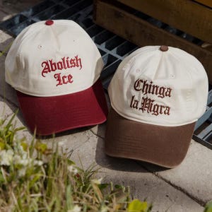 May include: Two baseball caps with white crowns and colored brims. One cap has a burgundy brim and the text "Abolish Ice" embroidered in red. The other cap has a brown brim and the text "Chinga la Migra" embroidered in brown.