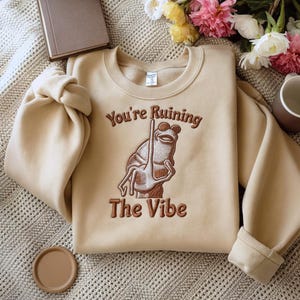 May include: Beige crewneck sweatshirt with the embroidered phrase "You're Ruining The Vibe" and a frog illustration. The frog is brown and holding a stick. The sweatshirt is folded on a textured surface.