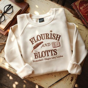 May include: Cream-colored sweatshirt with the embroidered text "FLOURISH AND BLOTTS" in brown, along with a feather and book graphic. The text "Booksellers - Diagon Alley, London" is also embroidered on the sweatshirt.
