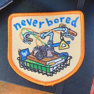 Pode incluir: Patch bordado com as palavras "never bored" em escrita azul. O patch apresenta um guiador de bicicleta, uma câmara, livros e um triângulo. O patch tem um fundo bege com uma borda laranja.