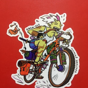 May include: A sticker featuring a cartoon goblin-girl riding an orange bicycle. The goblin-girl has yellow skin, a pink mohawk, and is wearing a white tank top. The bike has saddlebags and water bottles. Text on the sticker reads "goblin-girl" and "nomad patches".