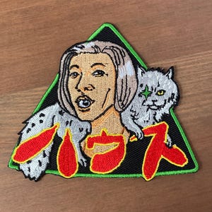 Puede incluir: Parche bordado con una mujer y un gato, sobre un triángulo verde. La mujer tiene el pelo rubio y una expresión de sorpresa. El gato es gris con una estrella. Texto japonés en rojo.