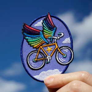 May include: A purple and blue embroidered patch of a bicycle with rainbow wings flying in a blue sky with white clouds.